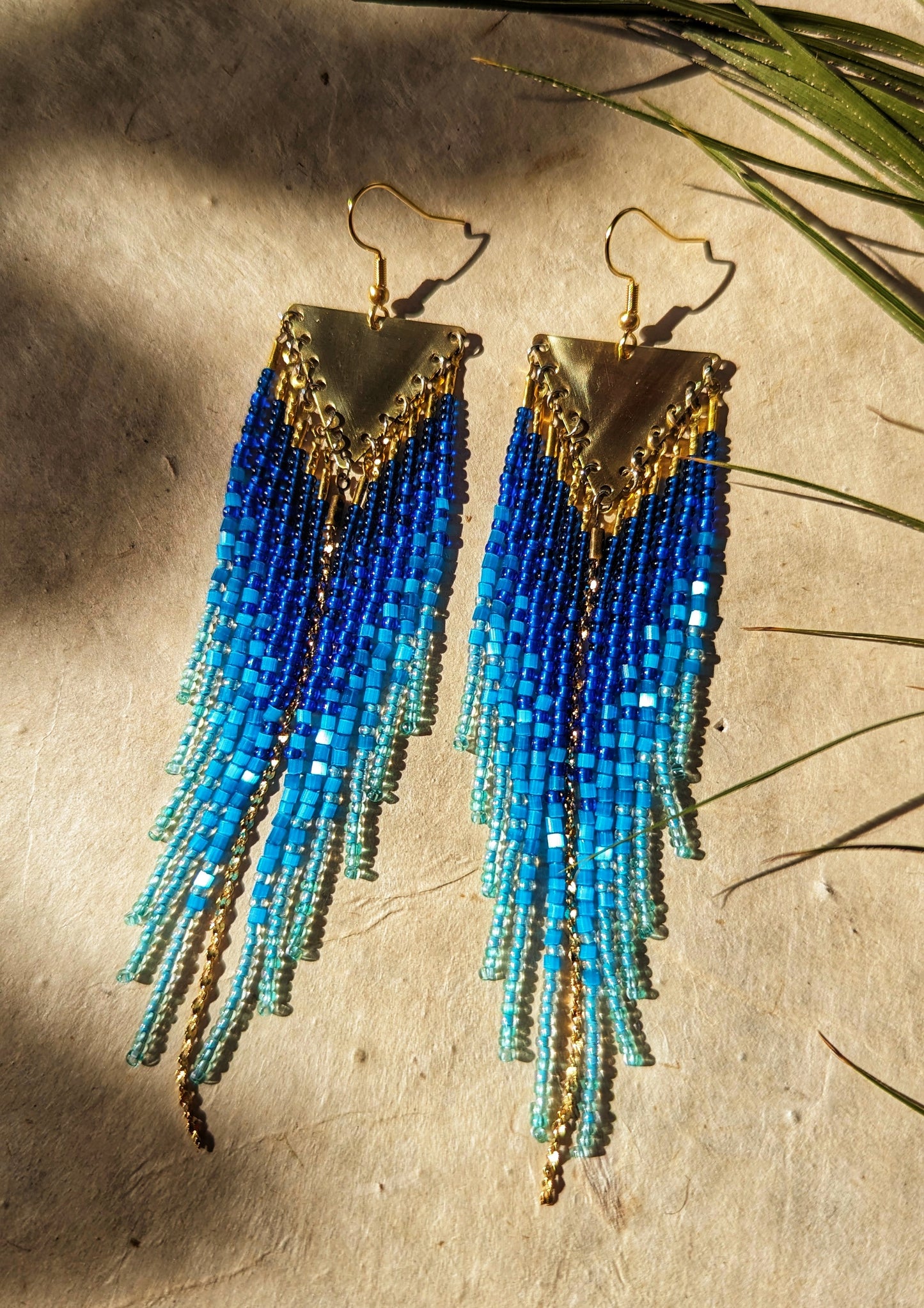 Seed bead earrings with blue and turquoise beaded strands in a gradient pattern. Triangular gold brass metal top.