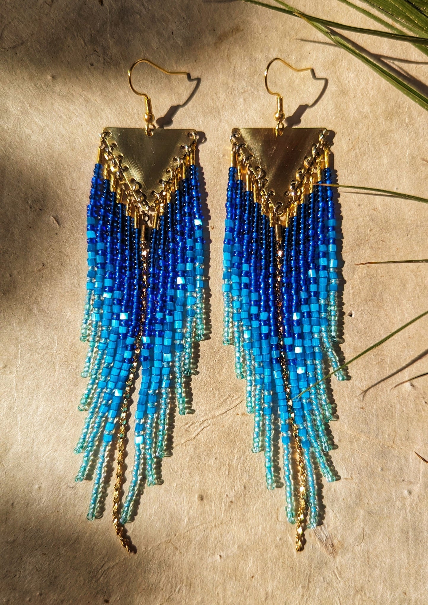 Seed bead earrings with blue and turquoise beaded strands in a gradient pattern. Triangular gold brass metal top.