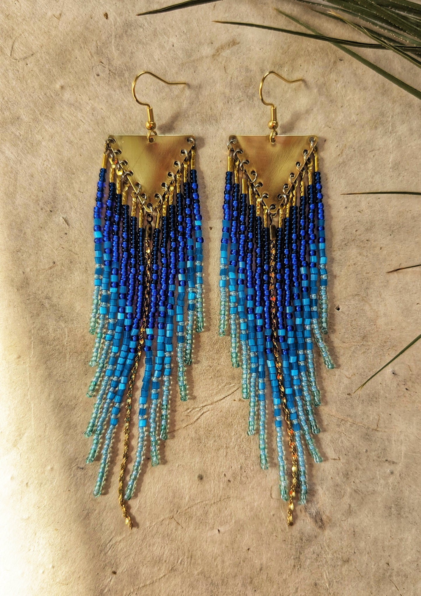 Seed bead earrings with blue and turquoise beaded strands in a gradient pattern. Triangular gold brass metal top.