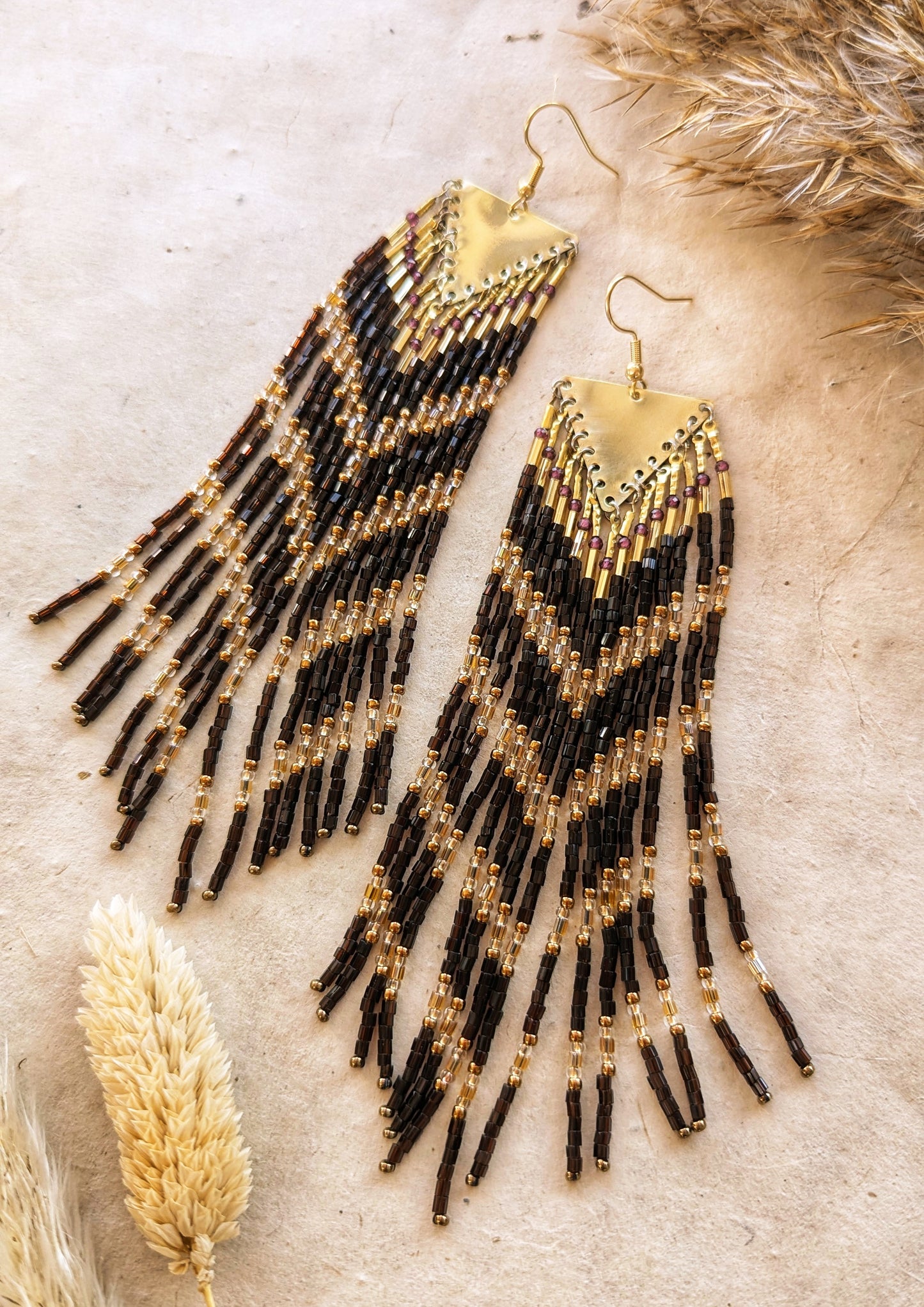 Moon & Milk - Long seed bead earrings with brown, gold, and bronze beads in a chevron pattern. Gold brass triangular tops. Photographed on a beige background with dried grasses.