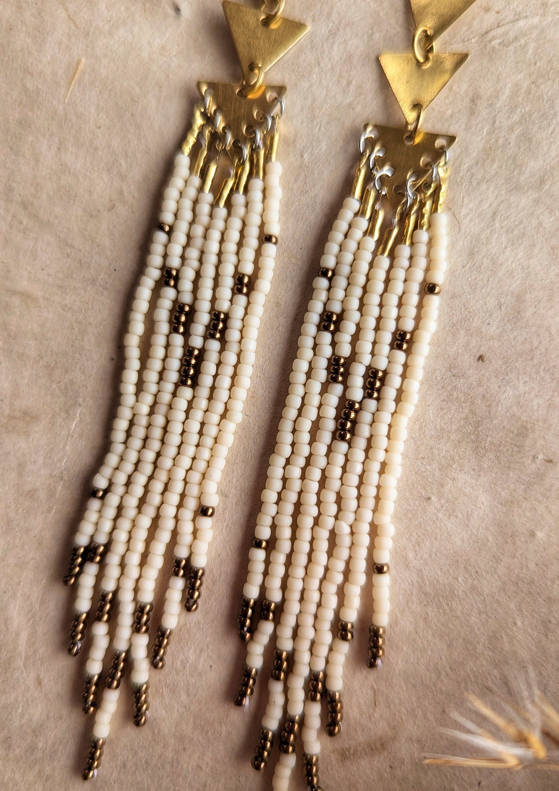 Moon & Milk - Gold triangle-link earrings with cascading cream and bronze seed bead strands, displayed on a neutral background with dried plants for decoration.