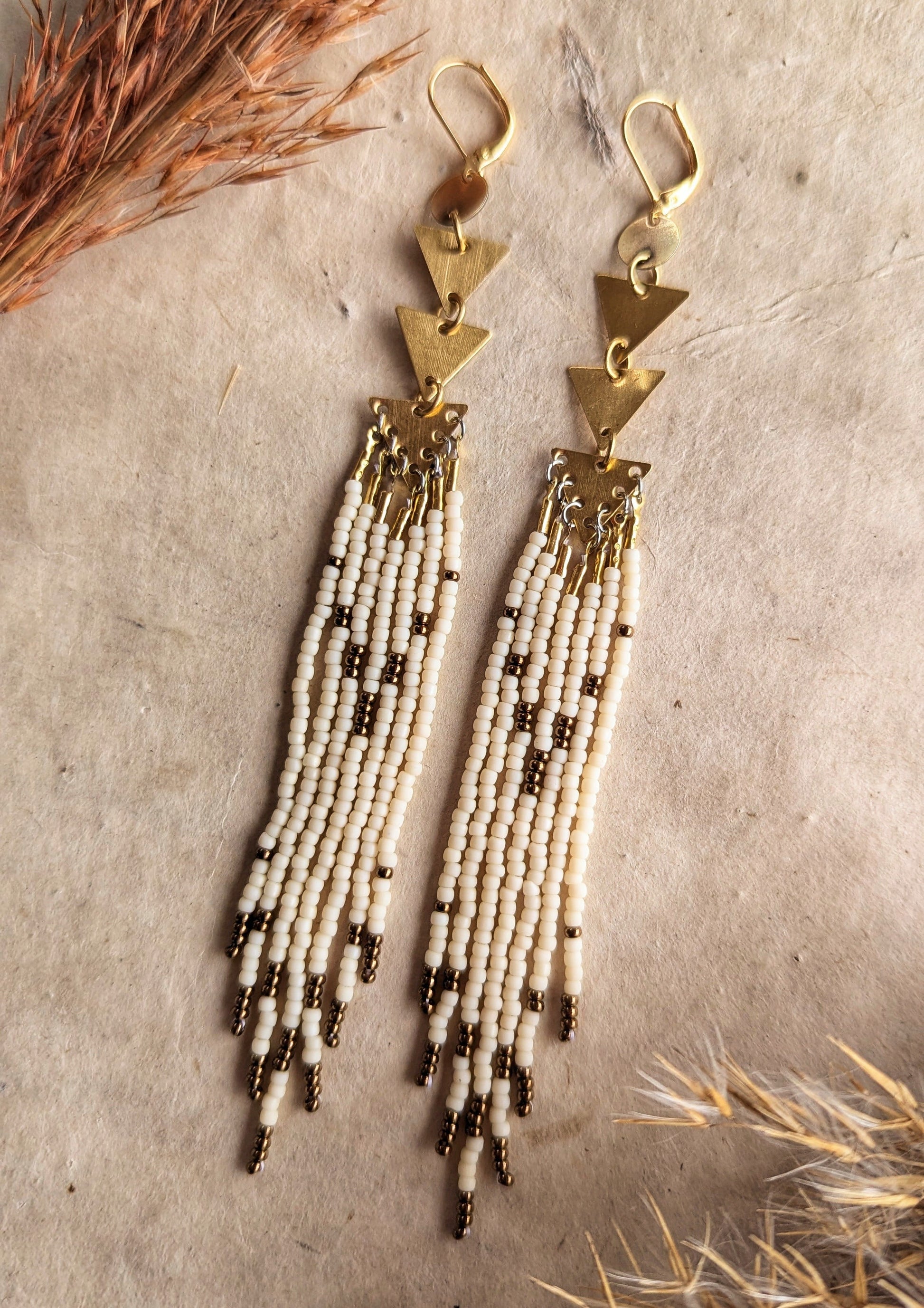 Moon & Milk - Gold triangle-link earrings with cascading cream and bronze seed bead strands, displayed on a neutral background with dried plants for decoration.