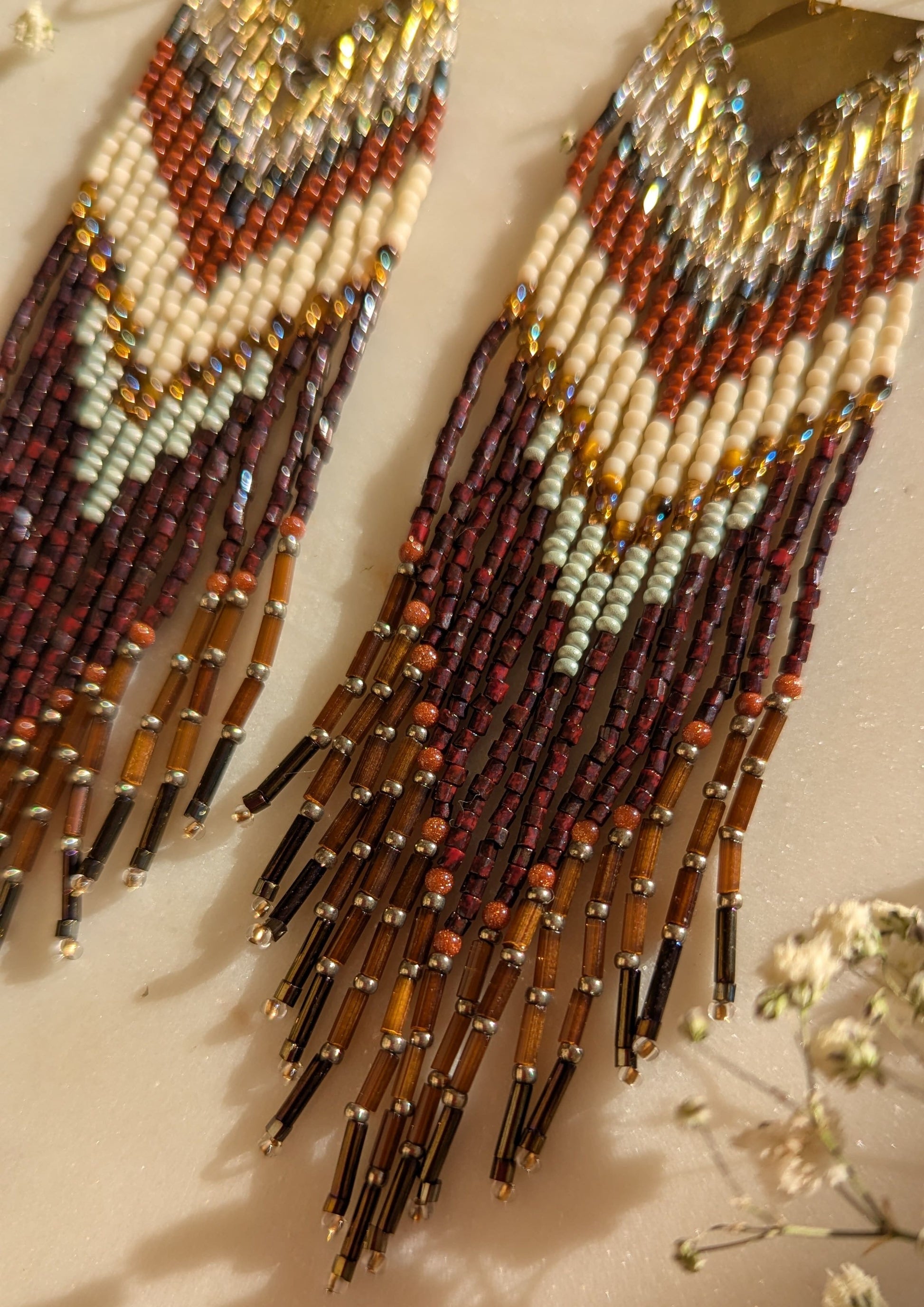 Handcrafted beaded fringe earrings with a chevron pattern in earthy tones of burgundy, gold, cream, and mint, accented by fringe with metallic gold and black details.