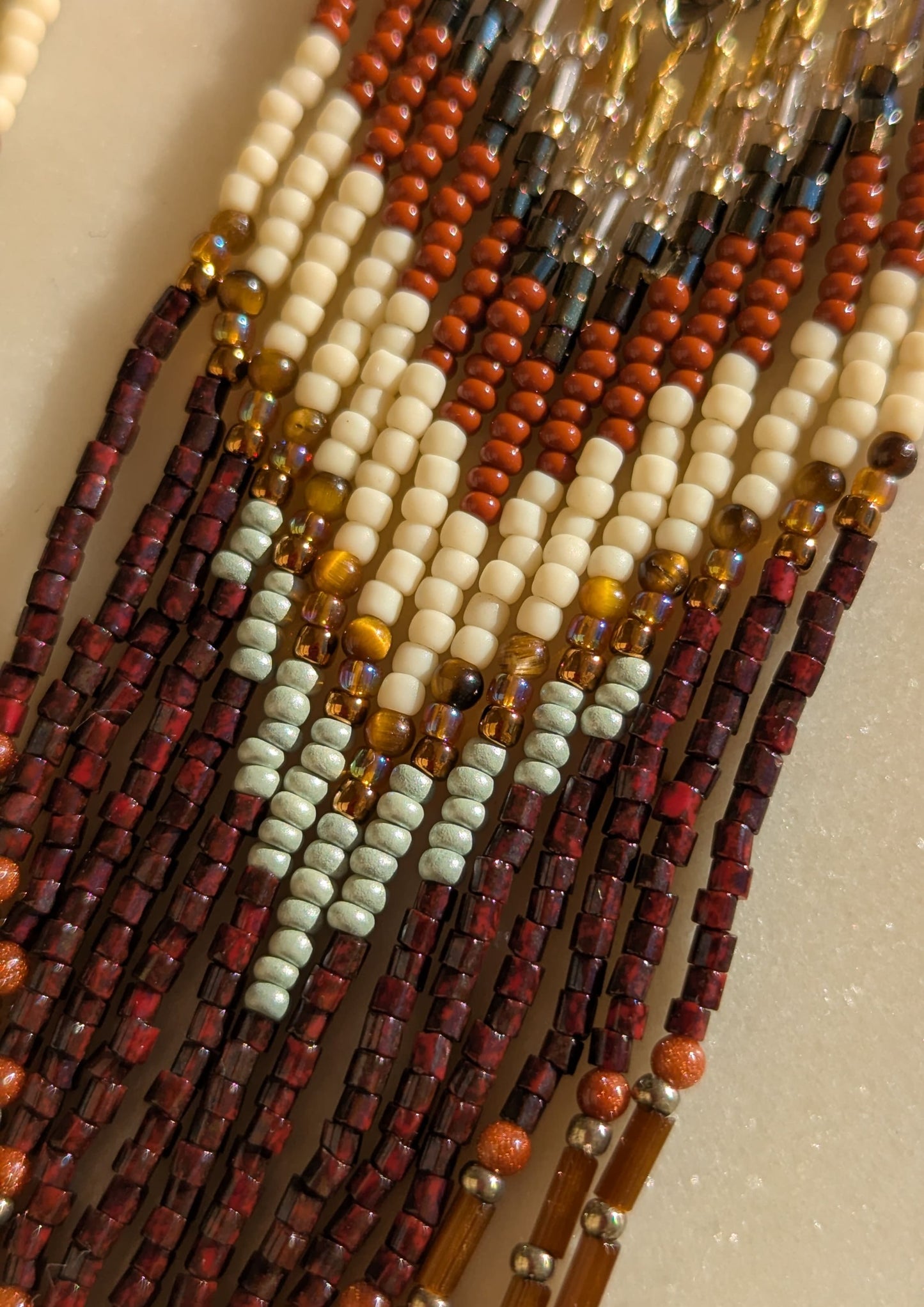 Handcrafted beaded fringe earrings with a chevron pattern in earthy tones of burgundy, gold, cream, and mint, accented by fringe with metallic gold and black details.