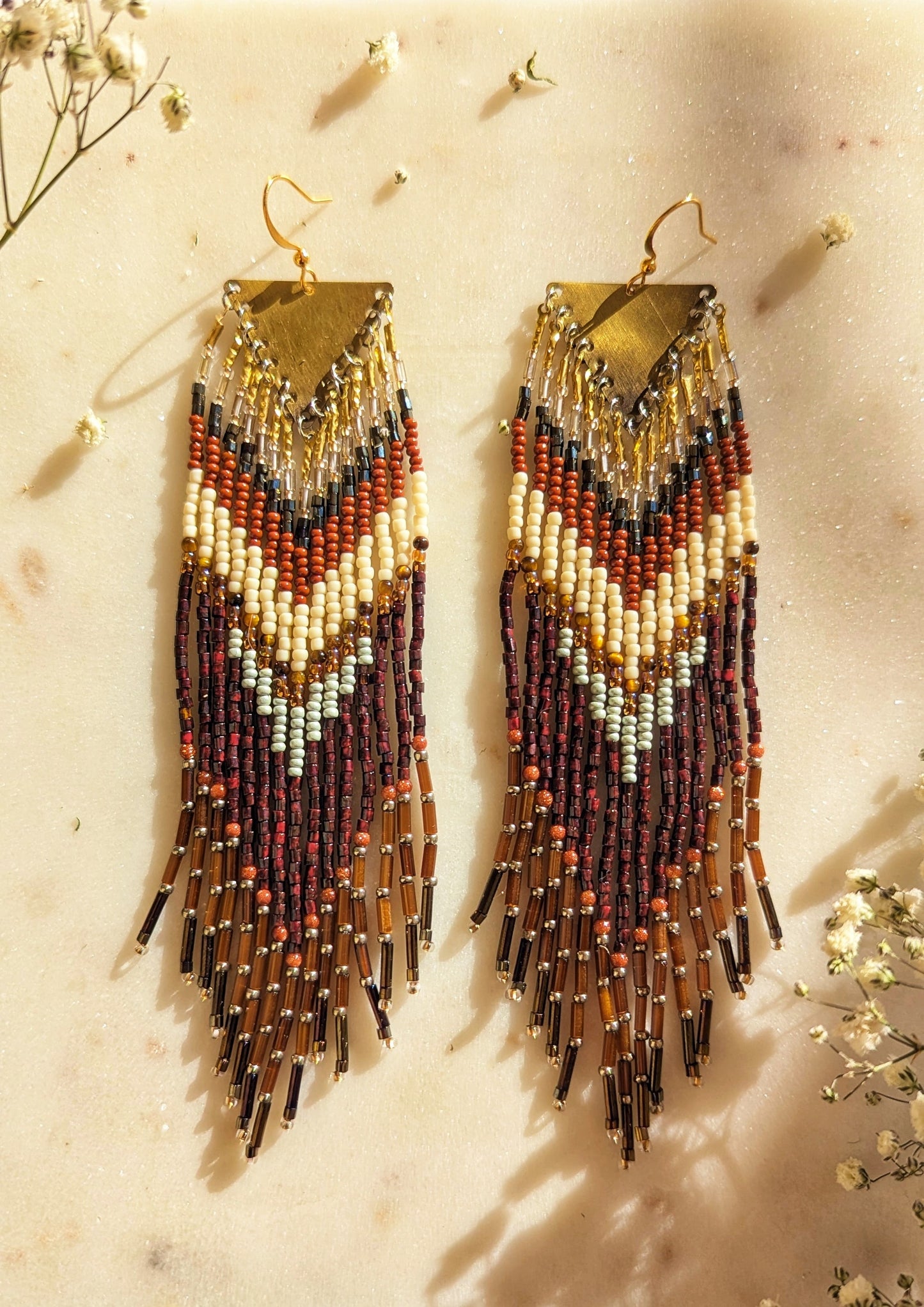 Handcrafted beaded fringe earrings with a chevron pattern in earthy tones of burgundy, gold, cream, and mint, accented by fringe with metallic gold and black details.