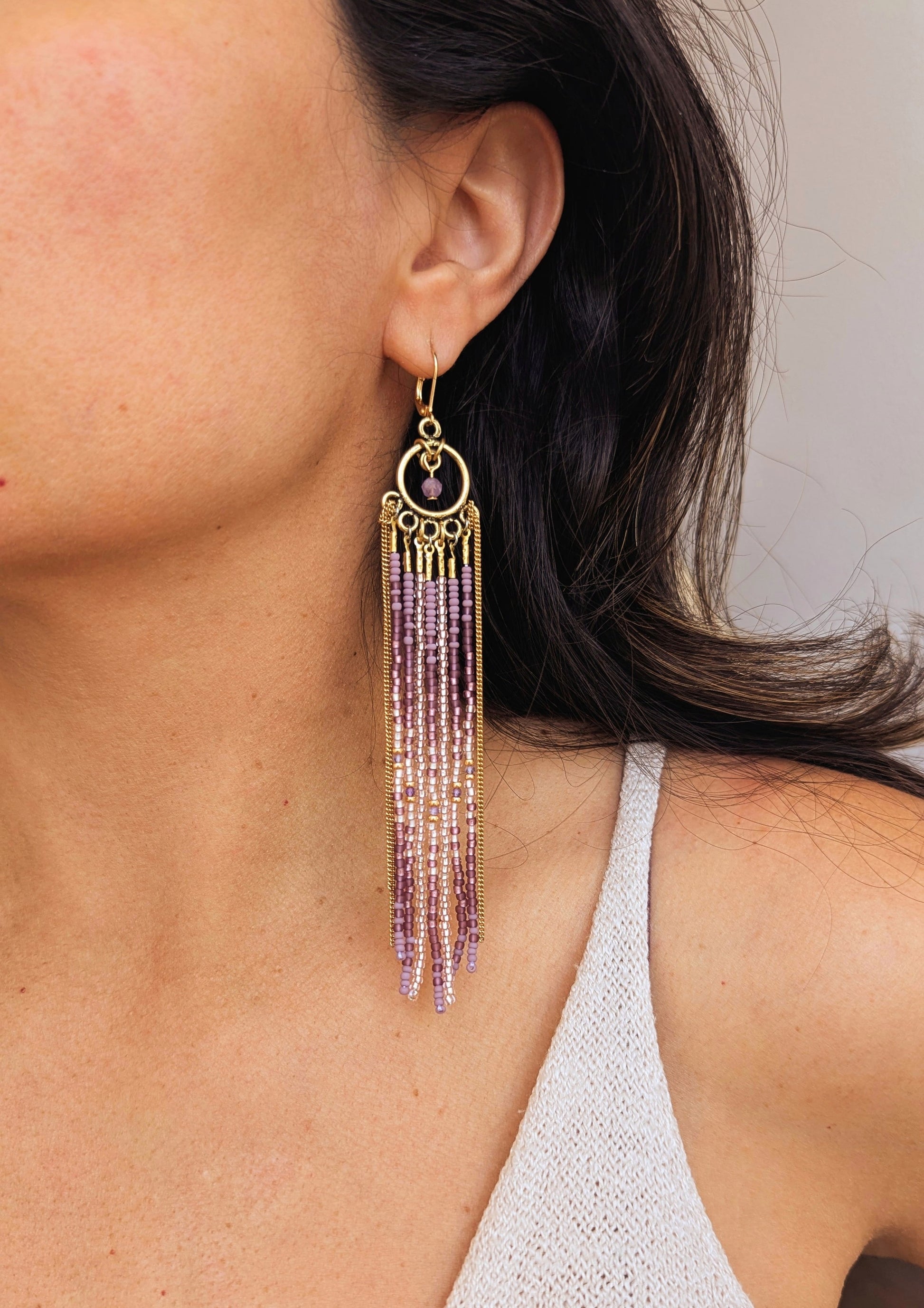 Moon & Milk handcrafted beaded earrings on a model, with glass beads in lilac and mauve tones, amethyst natural stones, accented by brass chain fringe in a long artisan earring design.