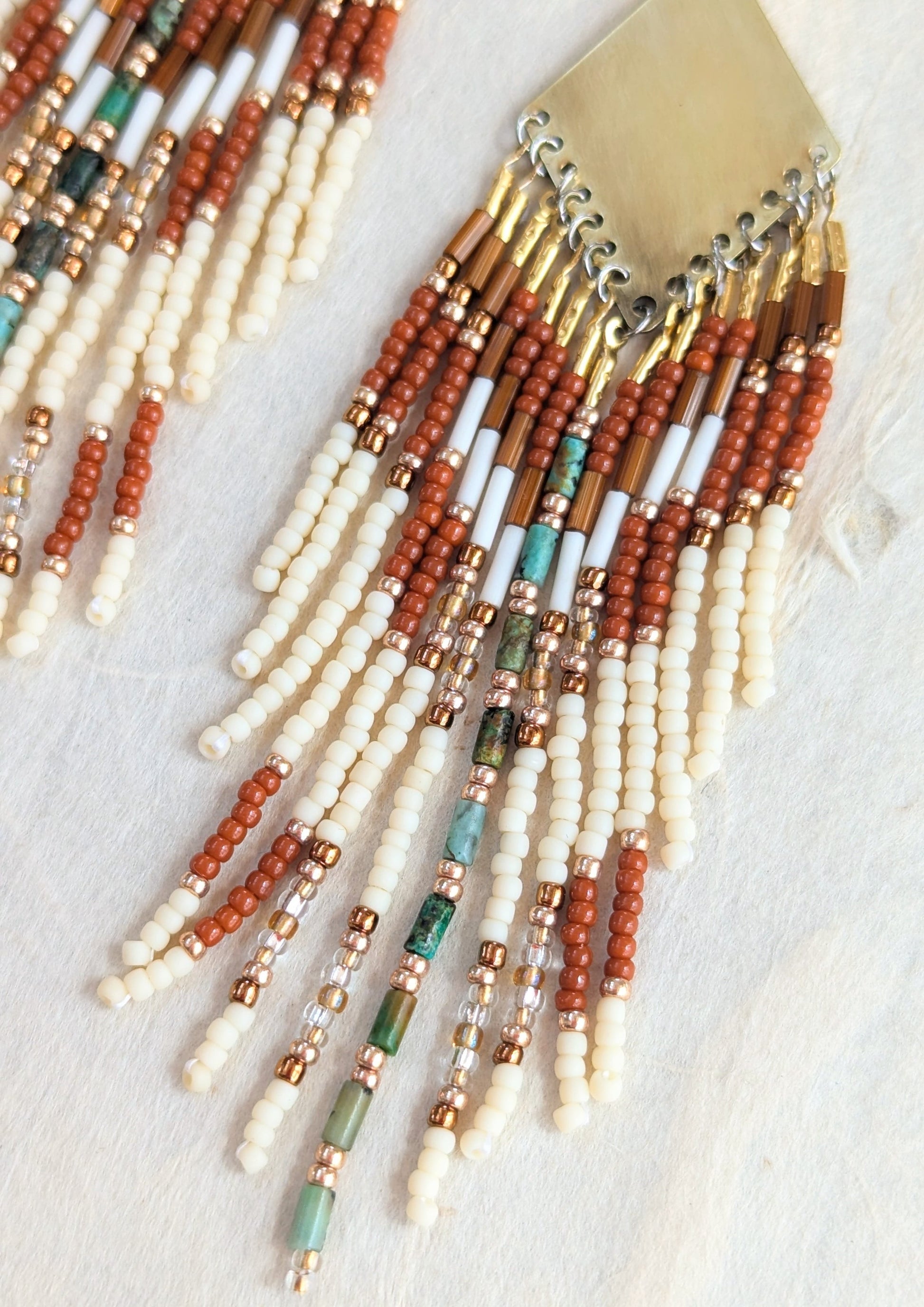 Handcrafted beaded earrings by Moon & Milk with brass diamond tops, rust and ivory seed beads, turquoise accents, and fringe design, photographed on linen fabric.