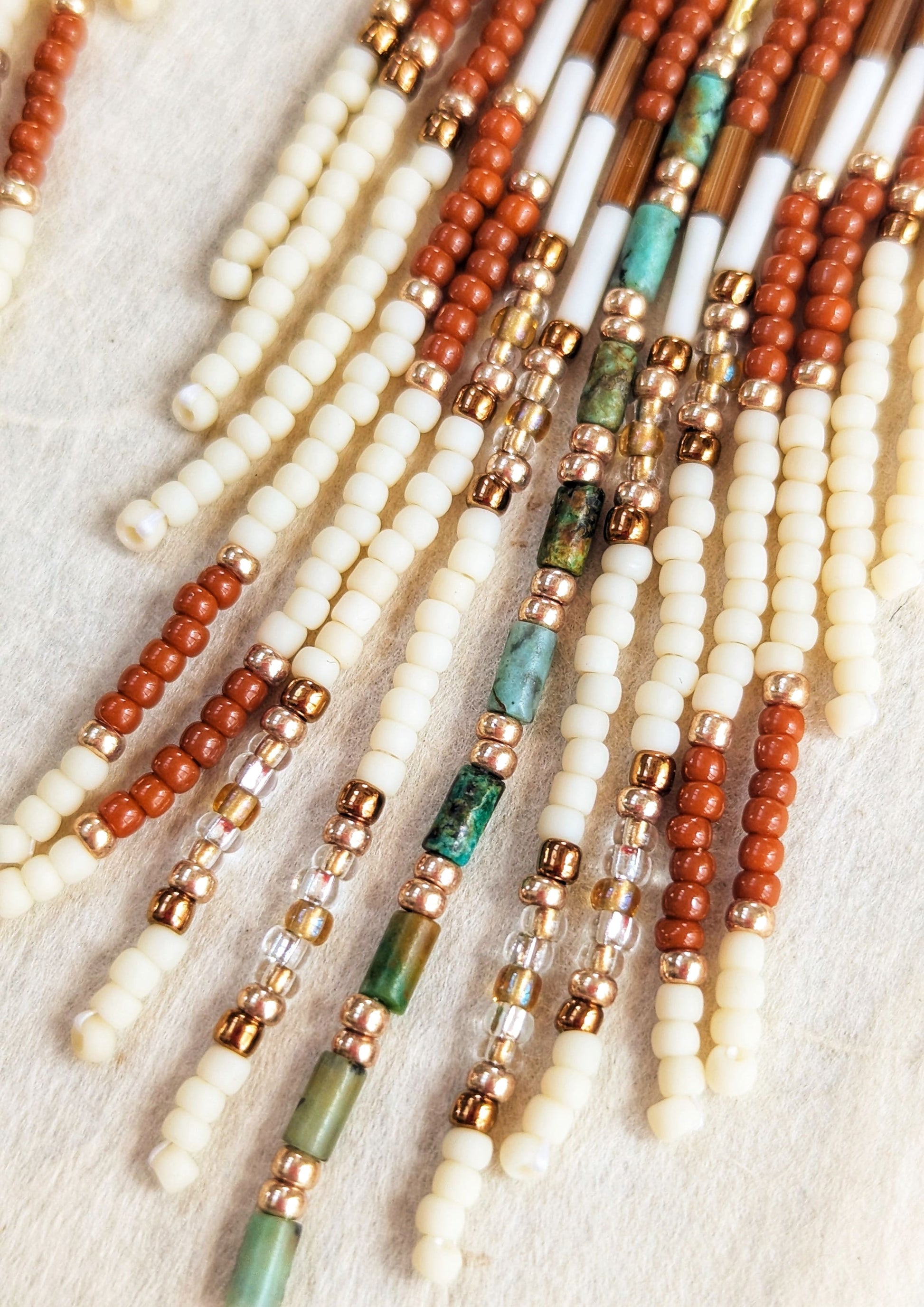 Handcrafted beaded earrings by Moon & Milk with brass diamond tops, rust and ivory seed beads, turquoise accents, and fringe design, photographed on linen fabric.