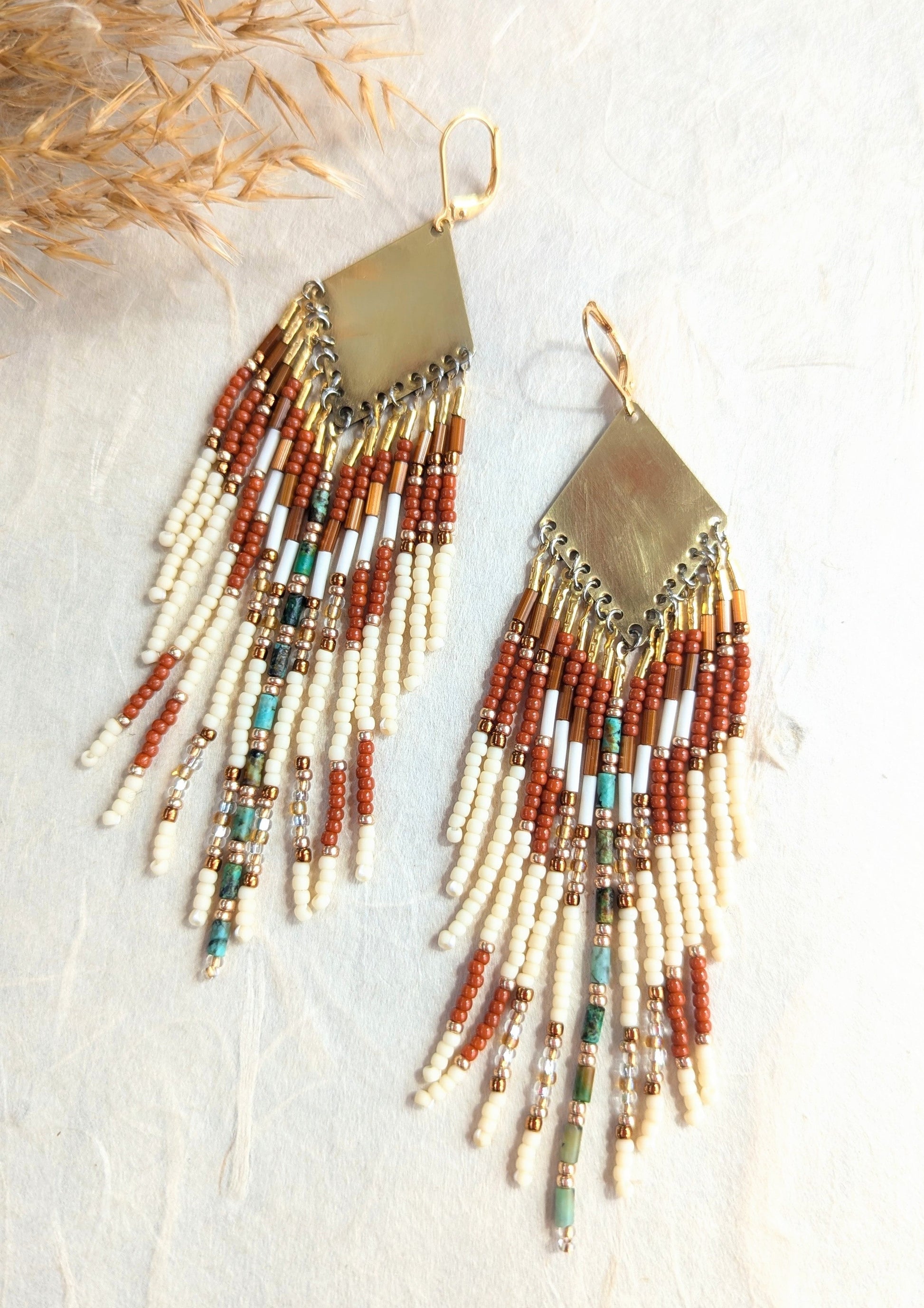 Handcrafted beaded earrings by Moon & Milk with brass diamond tops, rust and ivory seed beads, turquoise accents, and fringe design, photographed on linen fabric.
