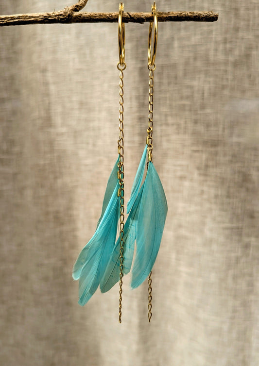 Turquoise feather earrings with gold-plated chain and hoops, hanging delicately against a neutral fabric backdrop.