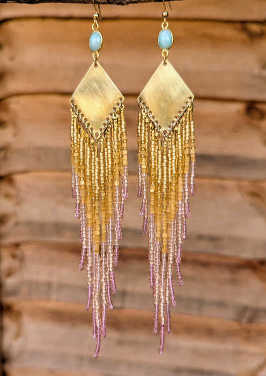 Long beaded earrings by Moon & Milk featuring a brass diamond-shaped base, light pink and gold seed bead fringe, and oval light blue stone studs.