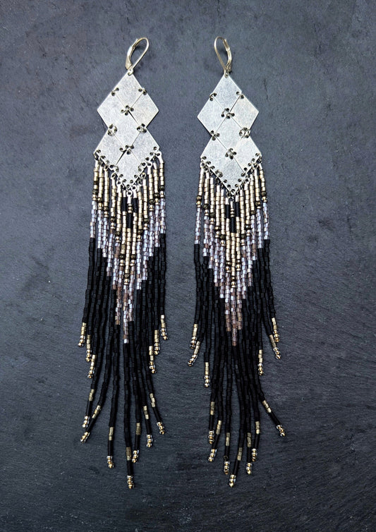 Pair of handcrafted beaded earrings with black, silver, and gold fringes on a dark slate background.