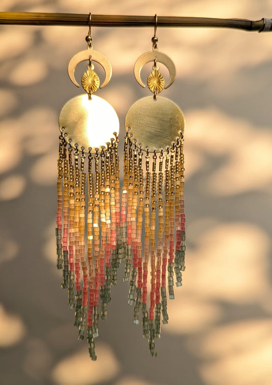 Gold sparkly earrings with beaded fringes, and brass moon findings against a blurred natural background.