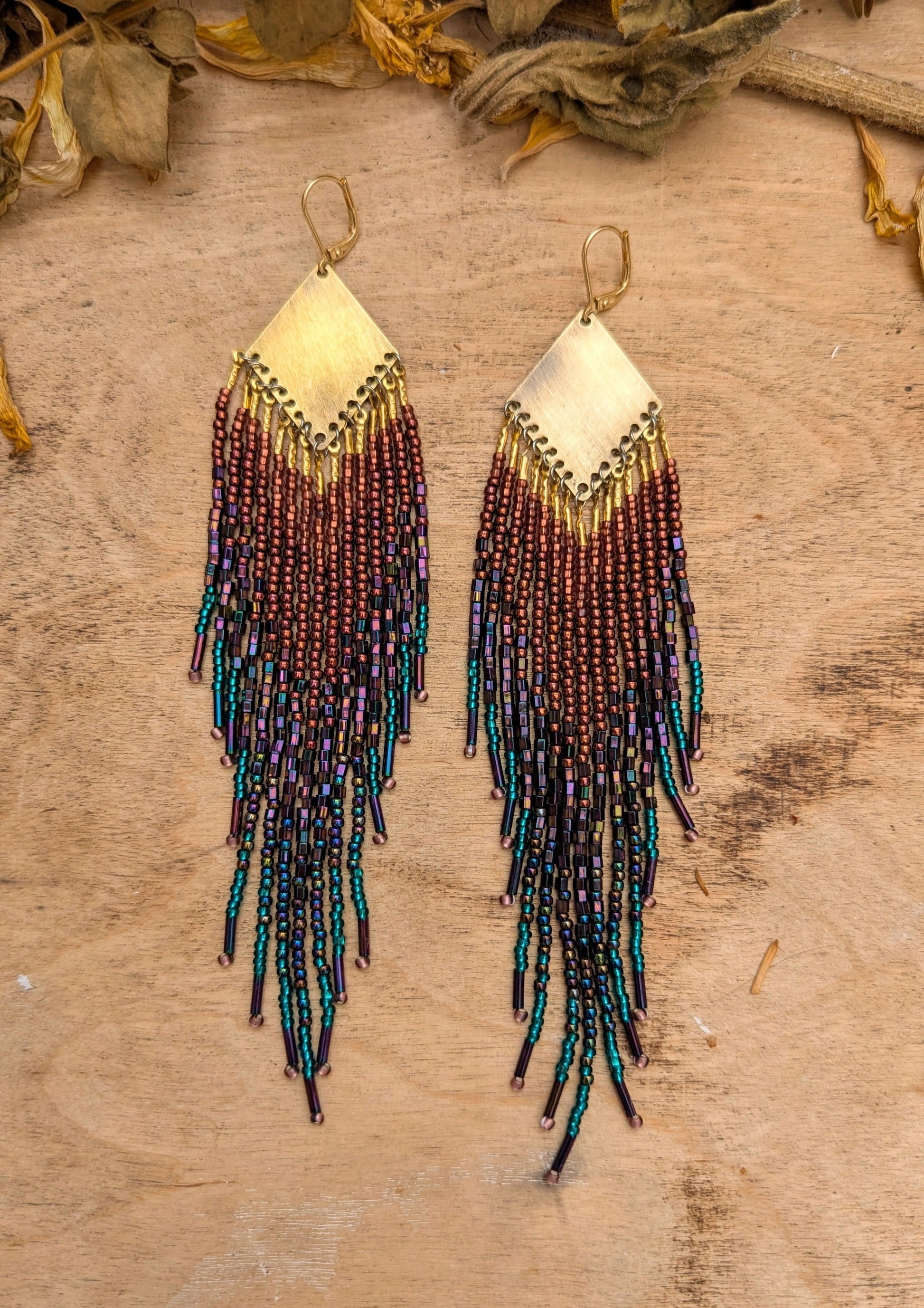 Hand-beaded fringe earrings on a wooden surface with dried leaves.