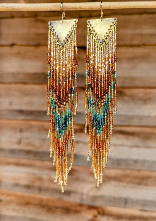 Seed beaded earrings with gold, brown, and teal beads hanging against a wooden background by Moon & Milk.