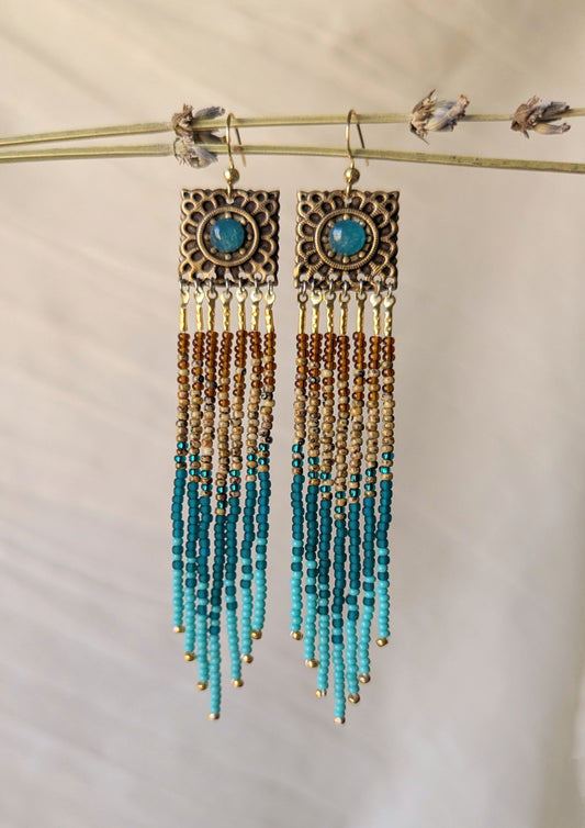 Pair of beaded earrings with turquoise and gold accents on a beige background by Moon & Milk.