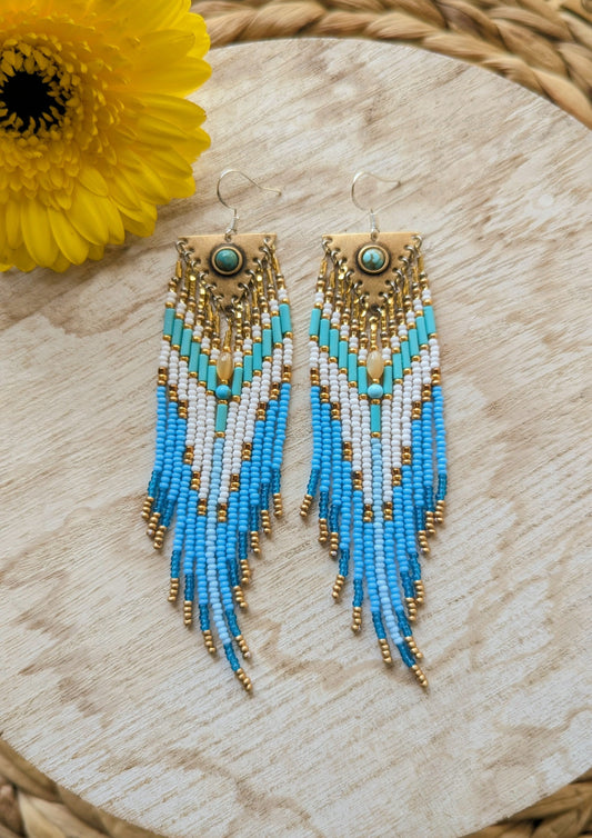 Beaded earrings with blue and gold fringes on a wooden surface with a yellow flower by Moon & Milk.