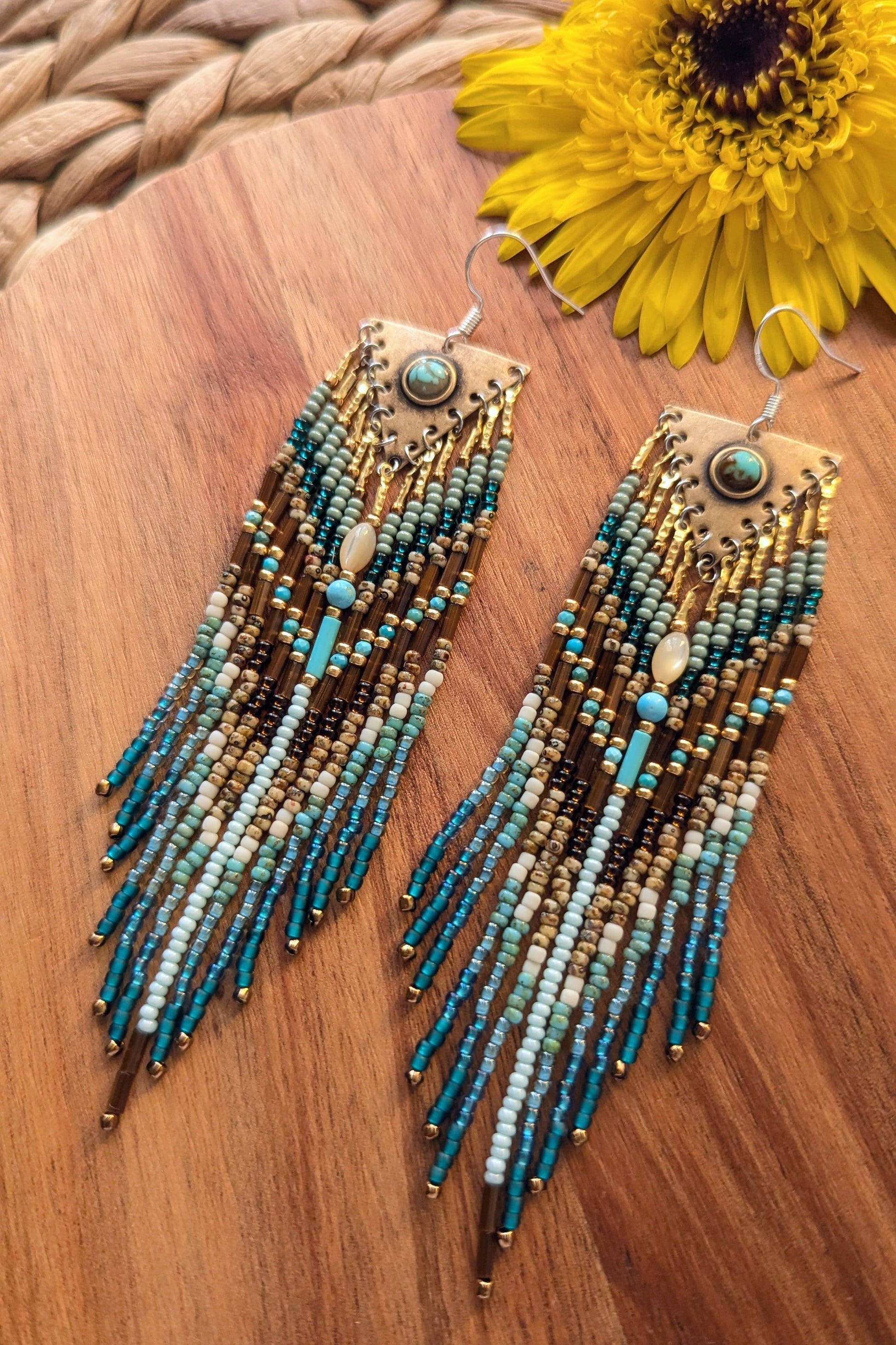 Handcrafted beaded earrings with teal and gold colors on a wooden surface with a yellow flower.