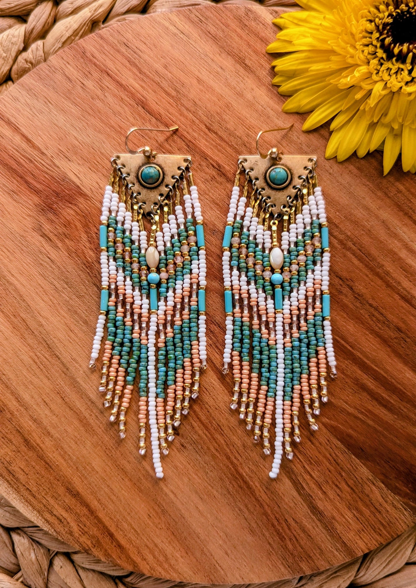 Beaded earrings with turquoise and peach pink accents on a wooden surface with a sunflower.