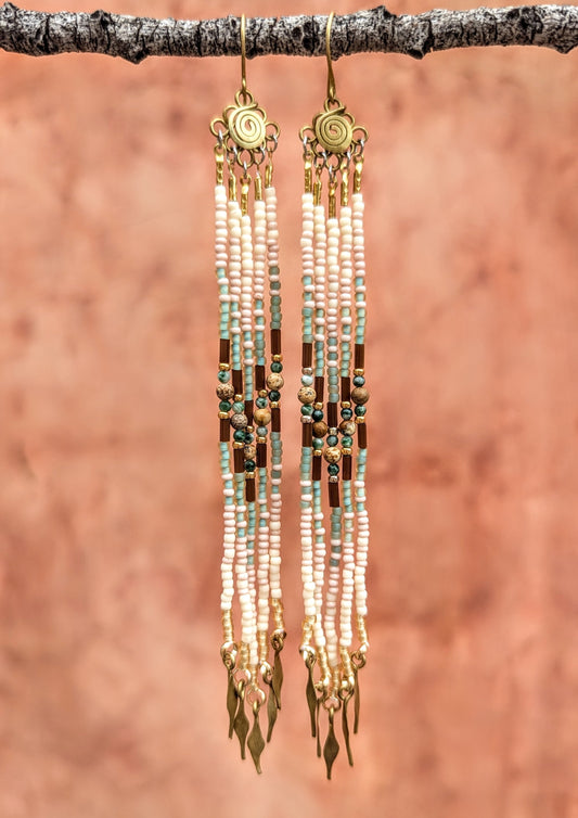 Long beaded earrings with gold accents hanging from a branch against a brown background.