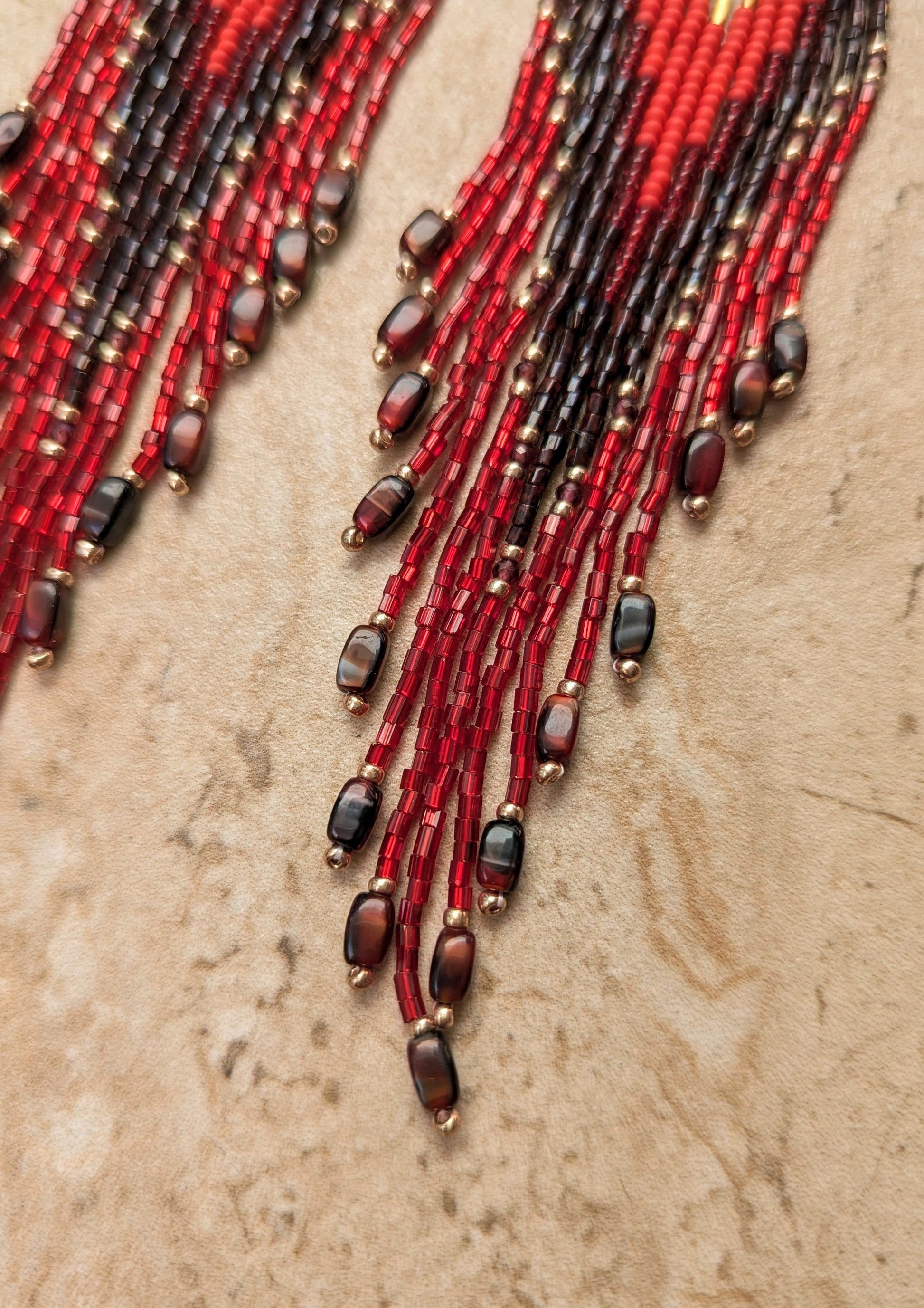 Bottom tips of beaded earrings with red, black, and gold beads on a textured beige surface.