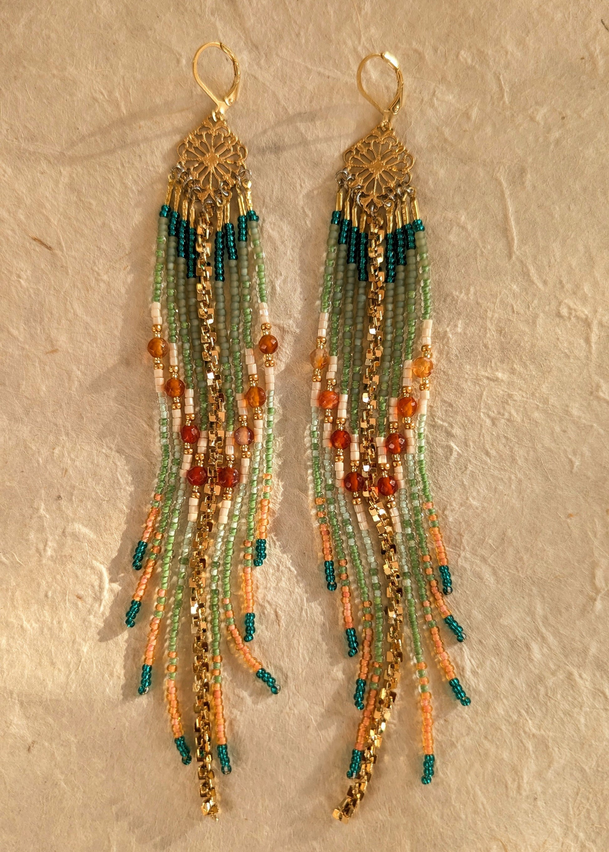 Pair of handmade beaded earrings with green, orange, and gold colors on a textured beige background.