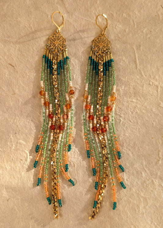 Pair of handmade beaded earrings with green, orange, and gold colors on a textured beige background.