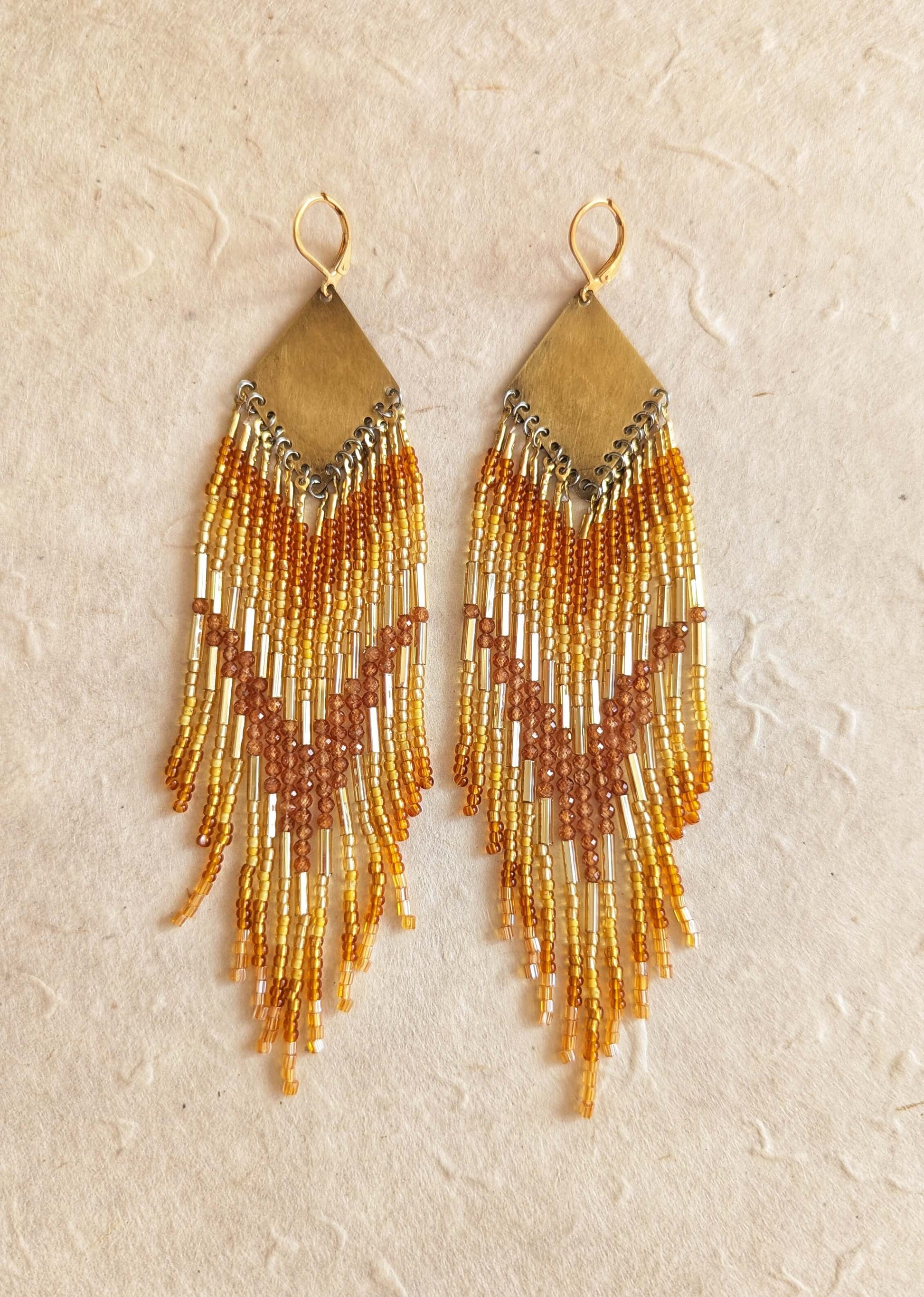 Pair of beaded earrings with gold bugles and hessonite garnet stones, and amber fringes on a beige background.