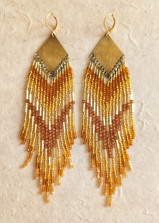 Pair of beaded earrings with gold bugles and hessonite garnet stones, and amber fringes on a beige background.
