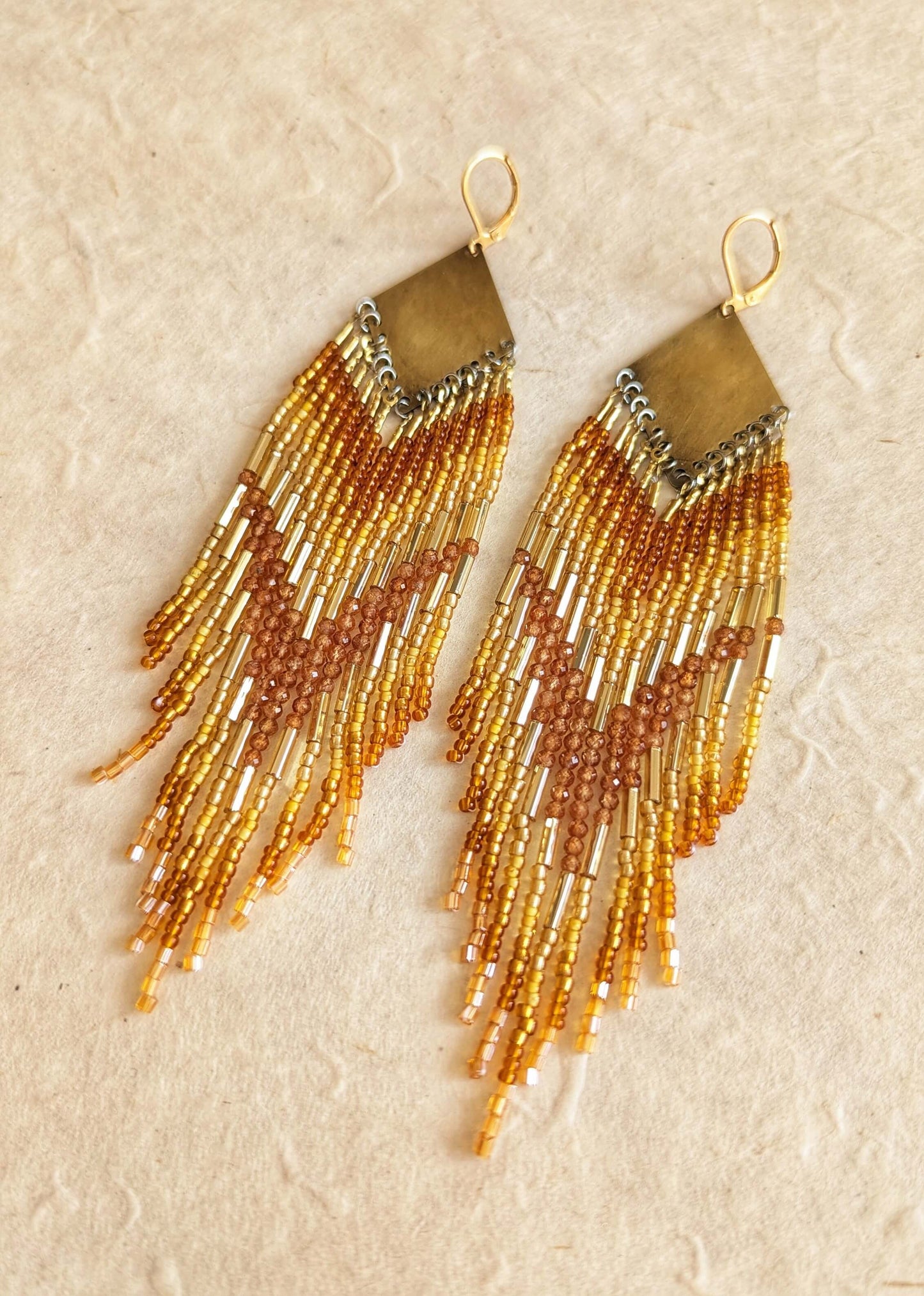 Pair of beaded earrings with gold bugles and hessonite garnet stones, and amber fringes on a beige background. By Moon & Milk.