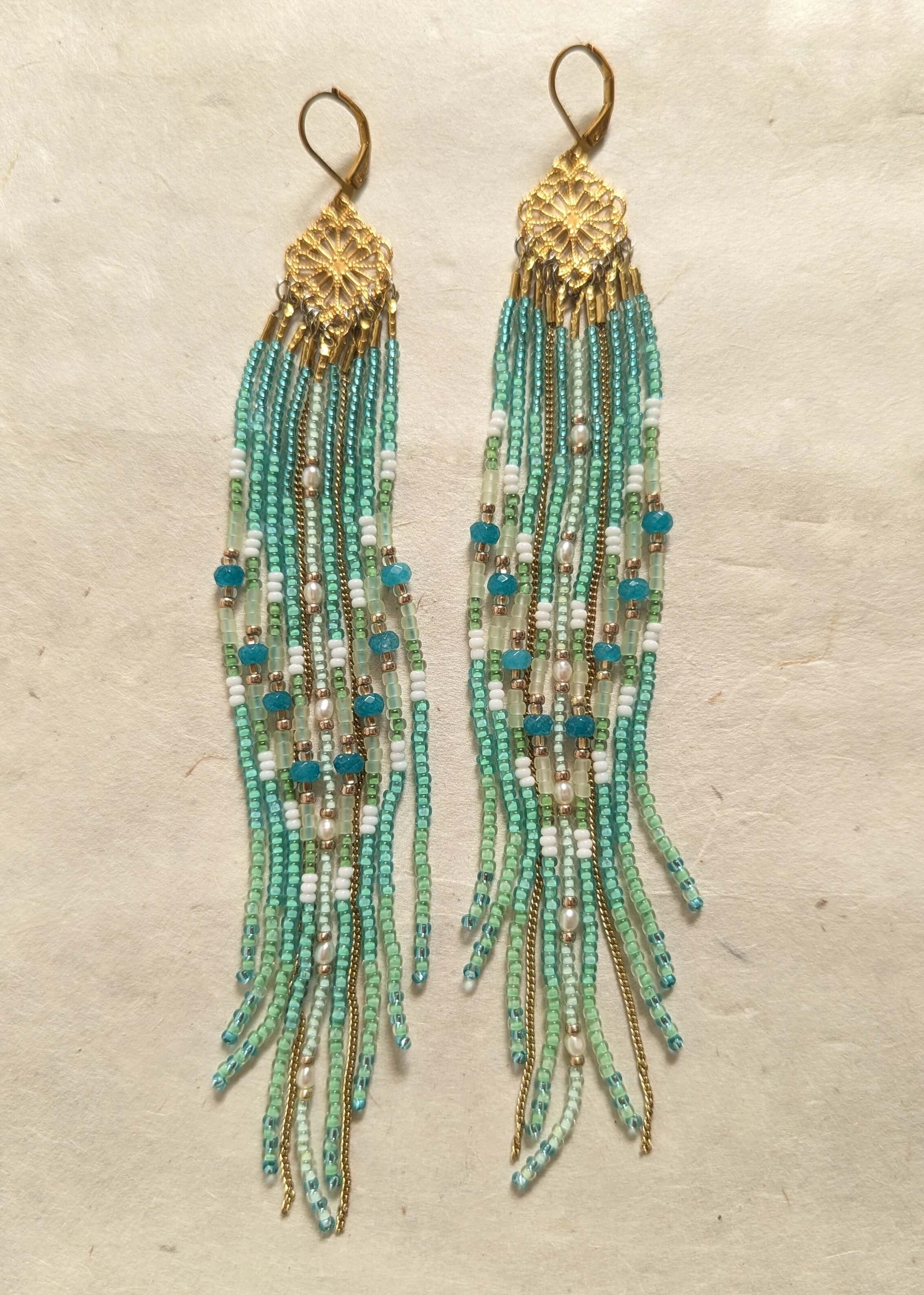 Beaded earrings with aqua, green, and gold fringes on a textured beige background.
