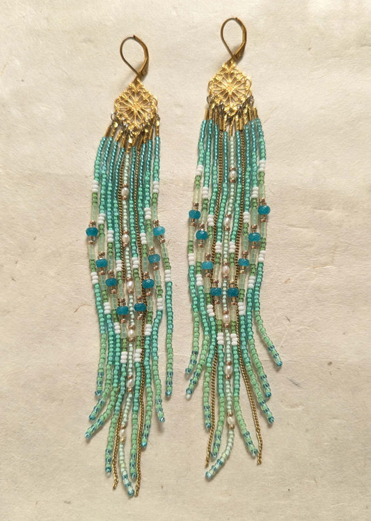 Beaded earrings with aqua, green, and gold fringes on a textured beige background.