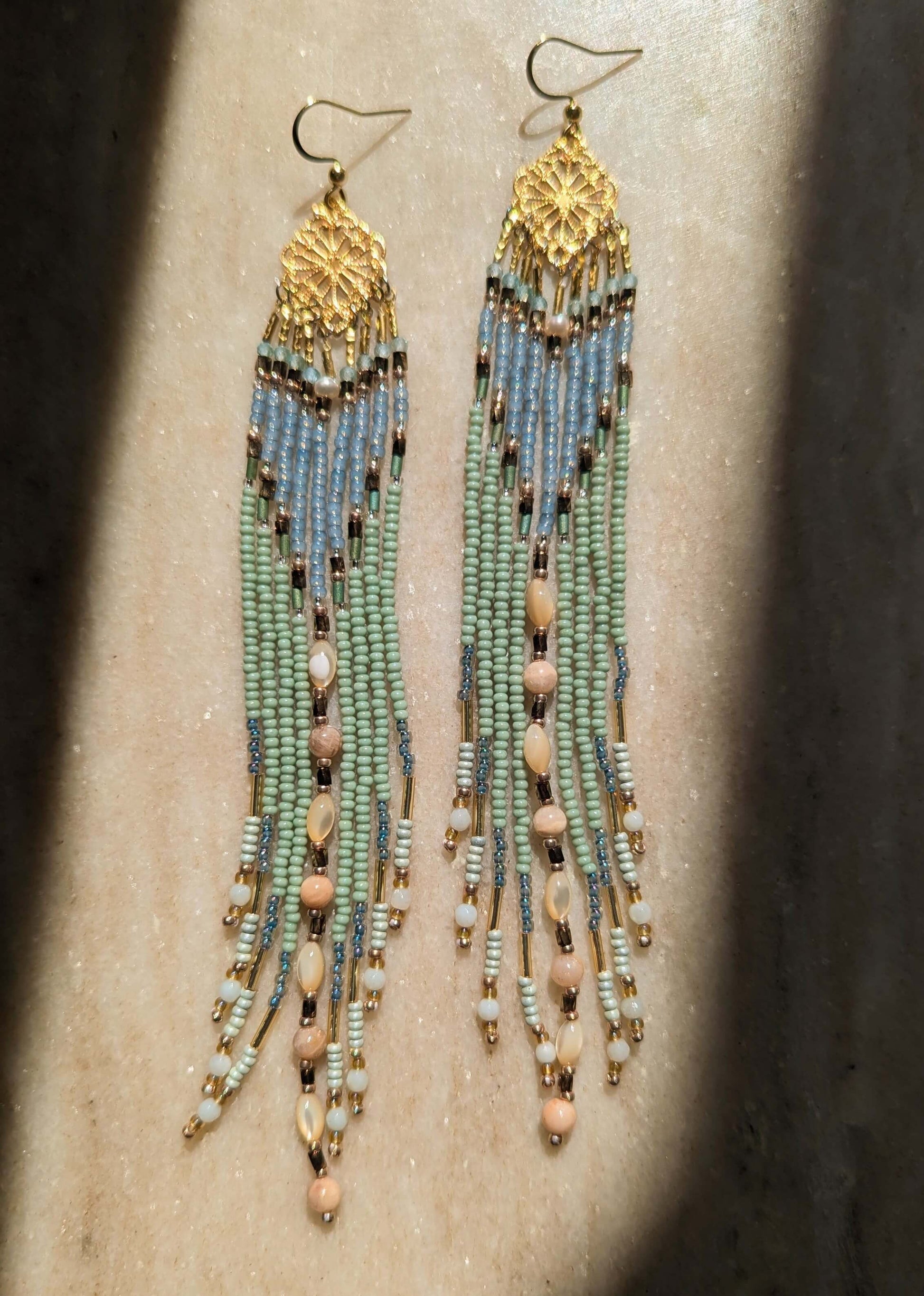 A pair of long beaded dangle earrings with gold-toned metal tops and hooks, featuring strands of light blue, mint green, and pale pink beads on a neutral surface.