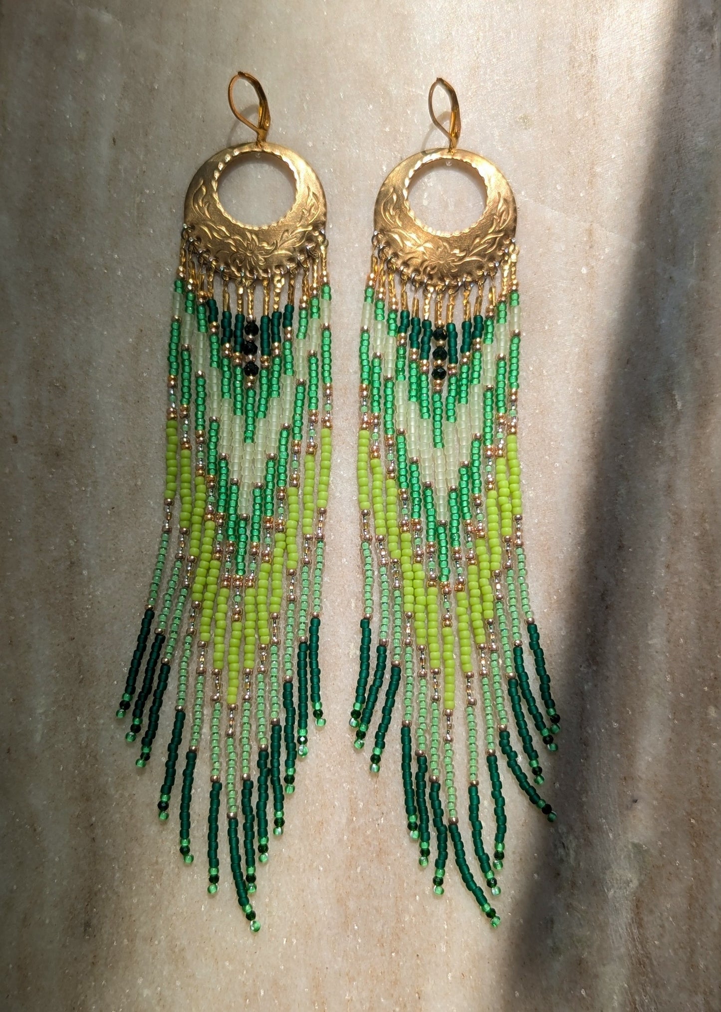 A pair of long dangling earrings with textured gold-tone circular tops and multiple strands of beads transitioning from light green to dark green, hanging against a neutral textured background.