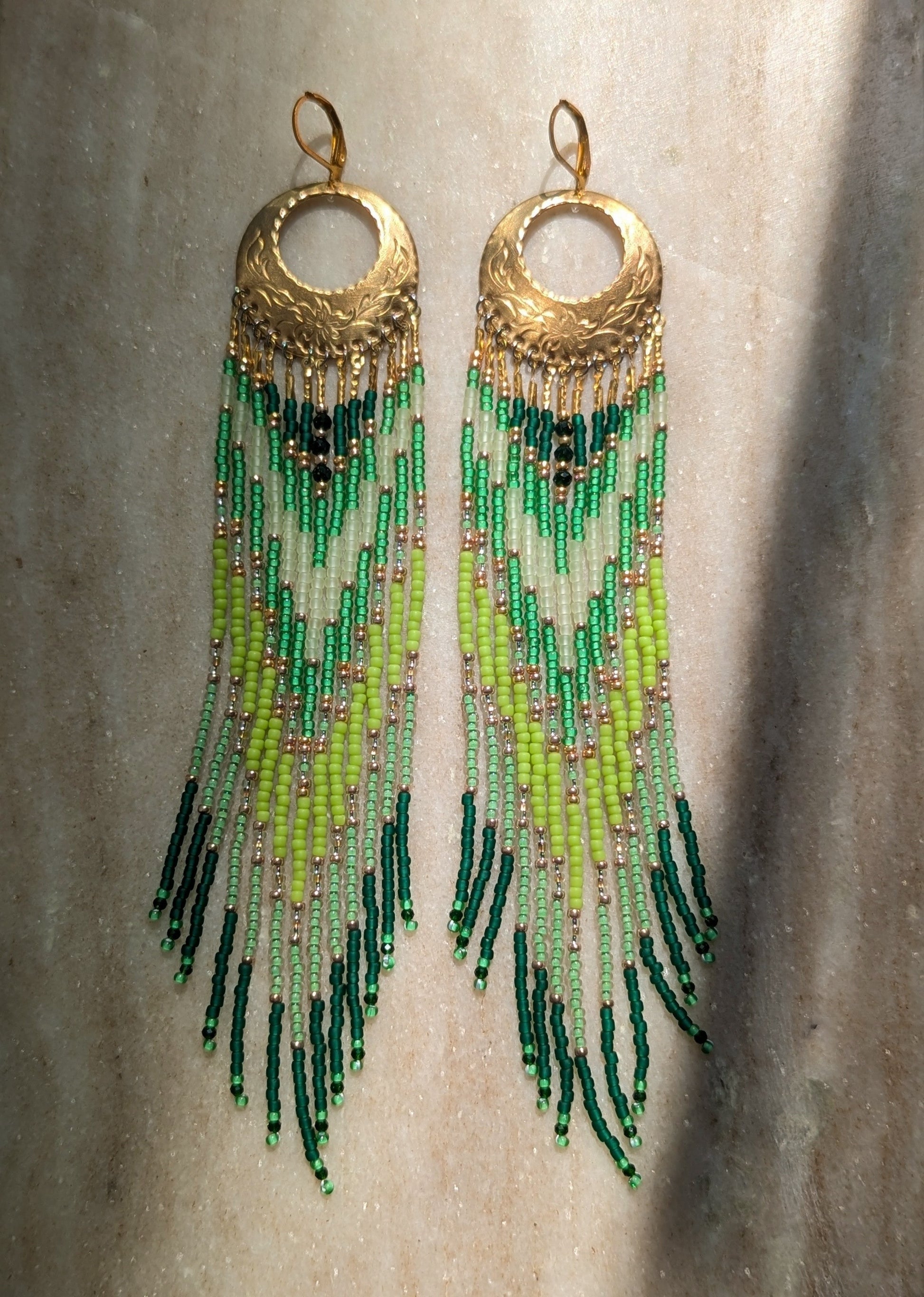 A pair of long dangling earrings with textured gold-tone circular tops and multiple strands of beads transitioning from light green to dark green, hanging against a neutral textured background.