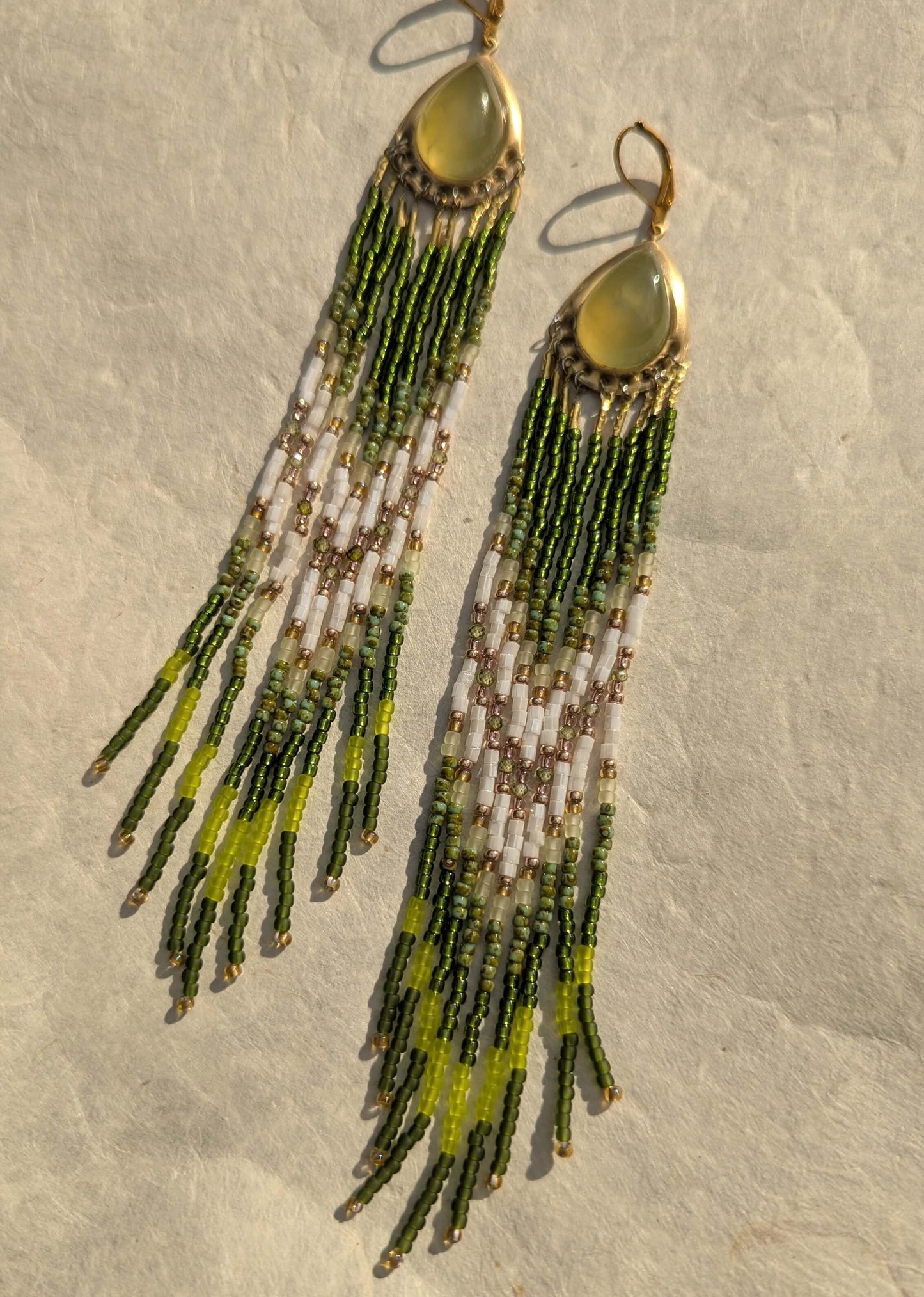 A pair of long beaded earrings with gold-tone hooks and oval gold tops, featuring green, white, and lime-yellow seed beads, displayed on a textured beige surface.
