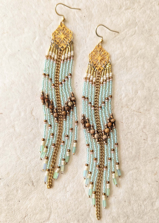 A pair of long beaded dangle earrings with gold-toned metal hooks and filigree tops, featuring strands of pale mint green, white, and brown seed beads on a textured beige background.