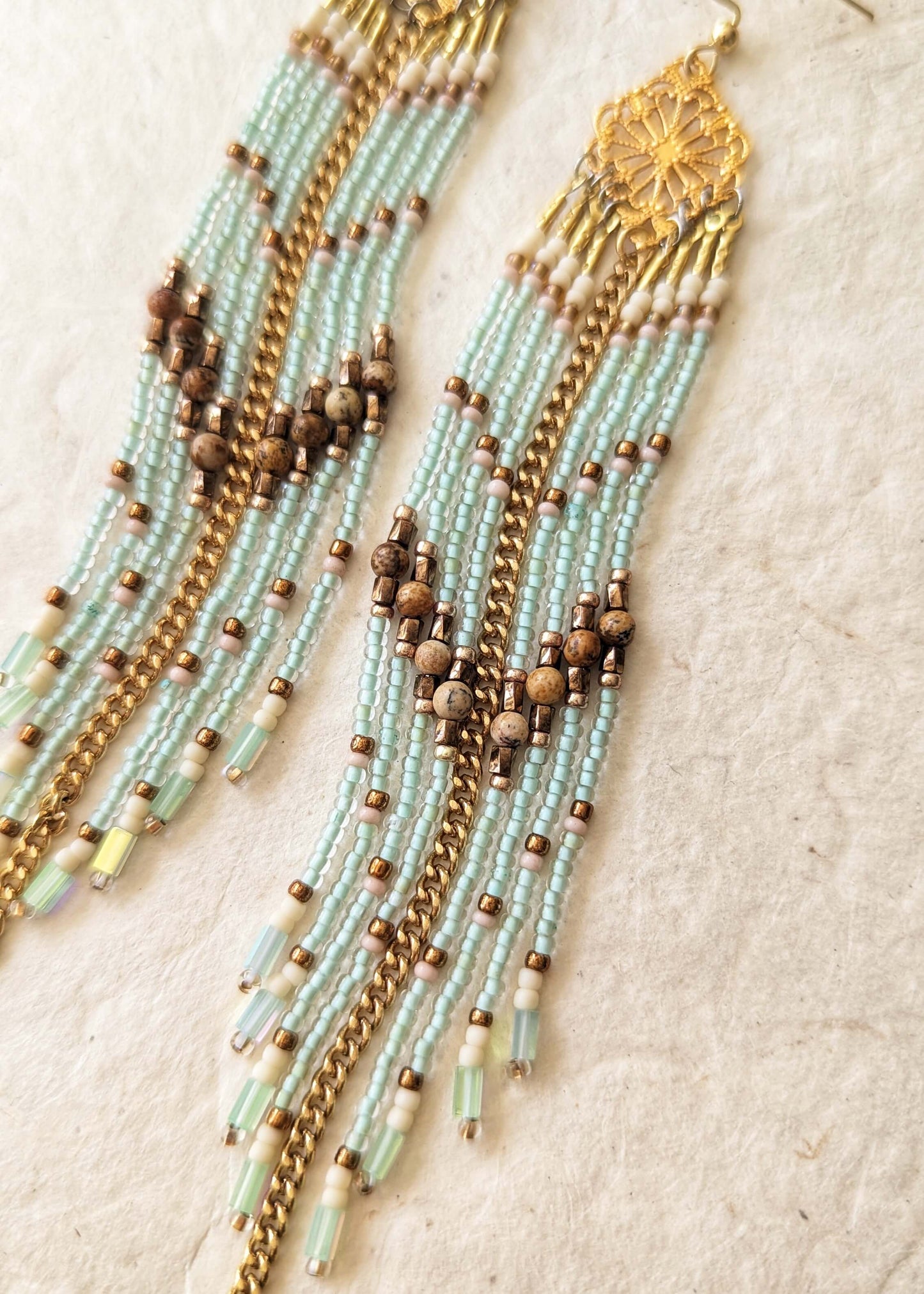 A pair of long beaded earrings with light blue beads, brown wooden beads, and gold-toned metal chains and charms, displayed on a textured beige fabric background.