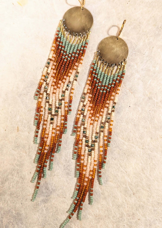 A pair of long beaded fringe earrings with round metallic gold-toned discs at the top, hanging from gold hooks. The dangling strands are made of small beads in tan, brown, cream, and light blue-green, arranged in multiple vertical strands.