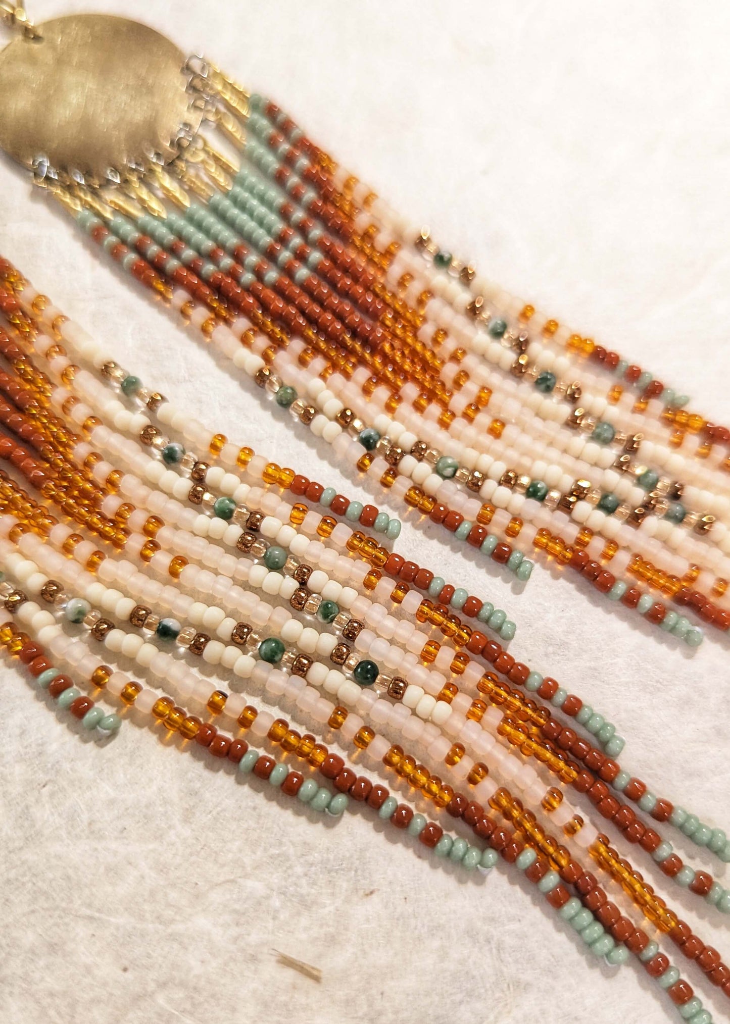 Close-up of beaded fringe earrings with multiple strands of cream, orange, brown, and pale blue-green beads, plus a gold-toned circular charm at the top, laid on a light textured surface.