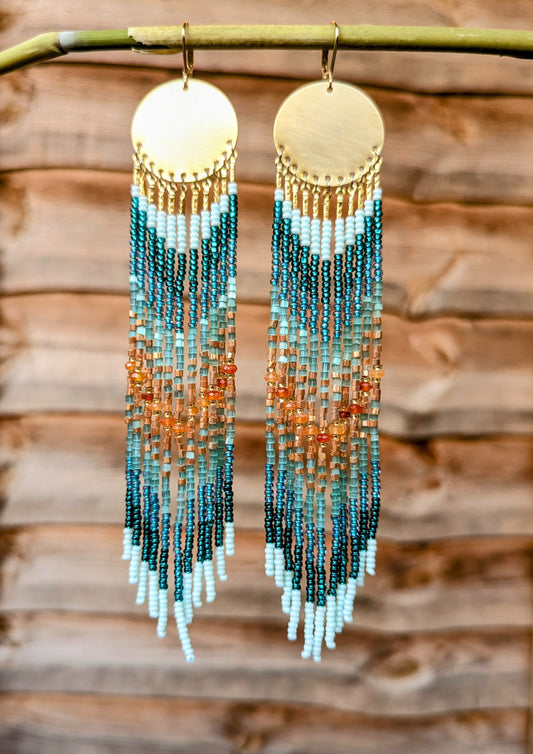 Hand beaded earrings with gradient colors hanging on a branch against a wooden background