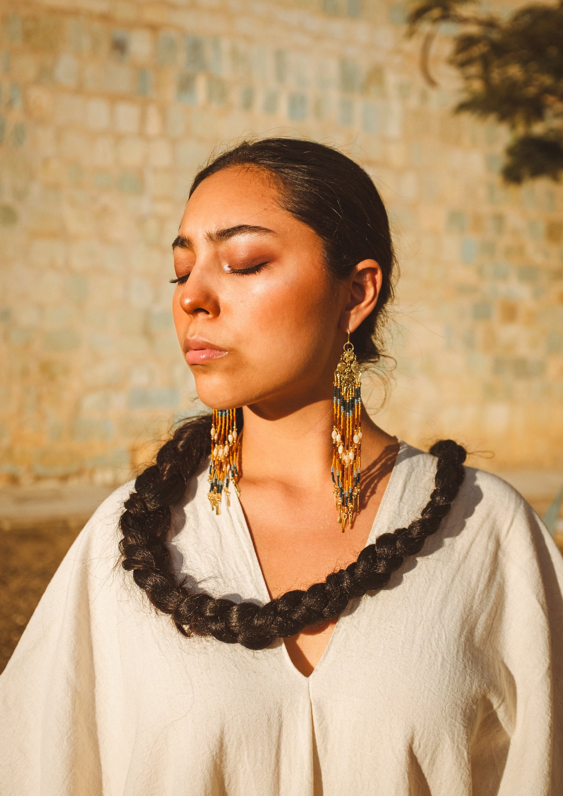 Woman with braided hair and gold beaded earrings by Moon & Milk against a stone wall.