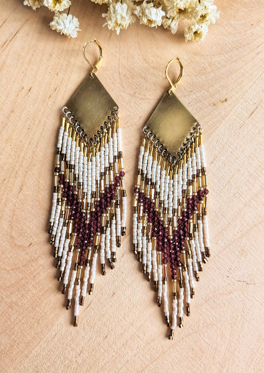 Long beaded earrings featuring garnet stones, ivory, and gold beads with brass diamond tops and chevron patterns, displayed on a wooden surface by Moon & Milk. 