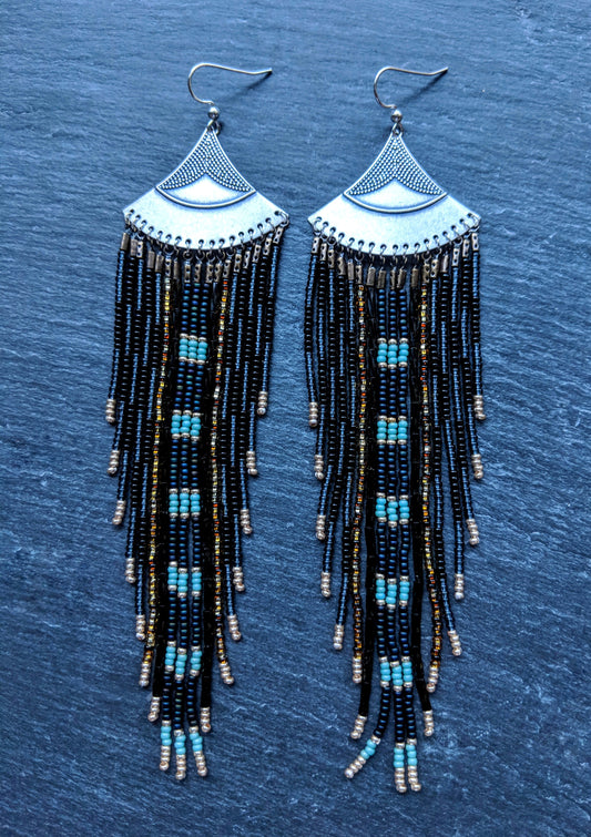 Moon & Milk long black and blue beaded earrings with silver fan tops and metallic accents, displayed on a slate-gray background.