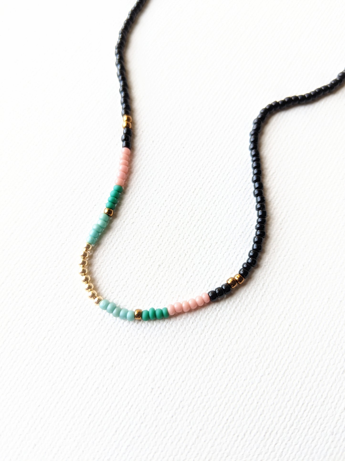 Minimalist beaded necklace with black, pink, green, and gold accent beads arranged on a white background, showcasing its clean and modern design.