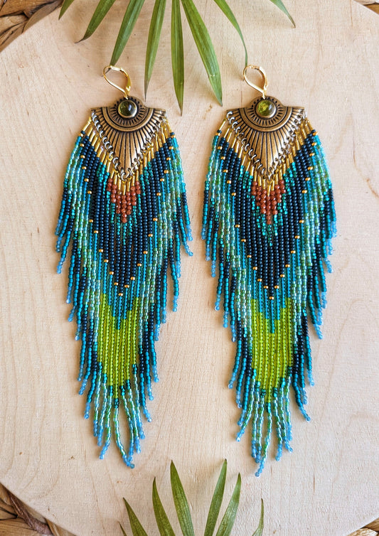 Pair of long beaded earrings featuring intricate patterns in deep blue, lime green, teal, and rust, with a brass sunburst accent at the top, and a dark green stone embedded, lying on a wooden surface.